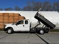 A white 2011 Ford F-350 SD with a black dump bed raised at an angle showing its dumping mechanism and a light bar on top