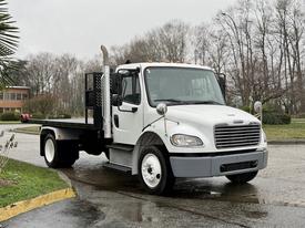 A 2019 Freightliner M2 flatbed truck with a white cabin and black flatbed features chrome mirrors and an exhaust stack on the side
