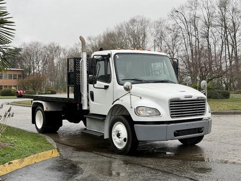 A 2019 Freightliner M2 flatbed truck with a white cabin and black flatbed features chrome mirrors and an exhaust stack on the side