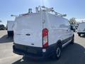 A white 2016 Ford Transit van with a ladder rack on the roof and a solid rear door parked in a lot