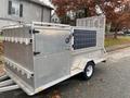 A silver 2023 Express Customs trailer with a side-mounted solar panel and multiple hooks along the sides