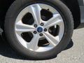 A close-up of a silver alloy wheel from a 2015 Ford Escape featuring five spokes and a central blue Ford logo with a black tire mounted on it