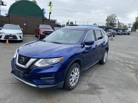 A blue 2019 Nissan Rogue is parked in a lot with its headlights on and a clean body design showcasing the front grille and alloy wheels