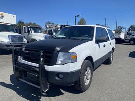 A 2013 Ford Expedition in white with a black hood and front grill guard parked with large trucks in the background