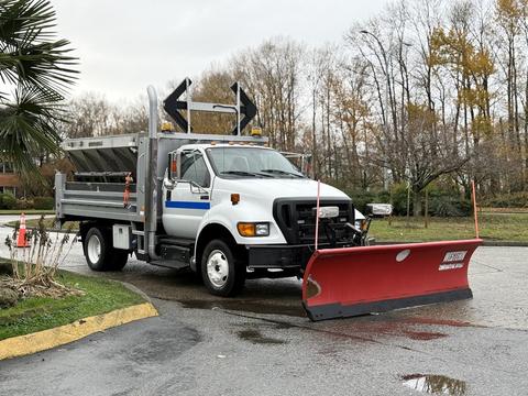 A 2013 Ford F-650 truck with a snow plow attached in front and a dump bed in the rear parked on a wet surface