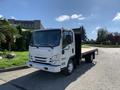 A 2015 Isuzu NPR truck with a flatbed design and a white exterior parked on a paved surface