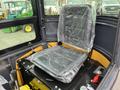 A black, padded operator seat in a mini excavator's cab partially covered with plastic wrapping, with control levers and safety features visible on the floor