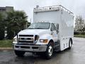 A white 2011 Ford F-650 box truck with a flat front and a roof-mounted light bar parked on a wet surface
