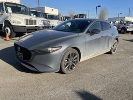 A 2023 Mazda MAZDA3 in gray with a slightly dirty exterior and black alloy wheels parked at a location with trucks in the background