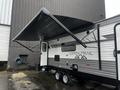 A 2022 Aspen Trail travel trailer with an extended awning and a door on the side featuring a simple design in black and white