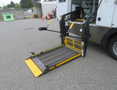 A wheelchair lift with a yellow and black platform extending from a van's rear, featuring handles for operation and a textured surface for traction