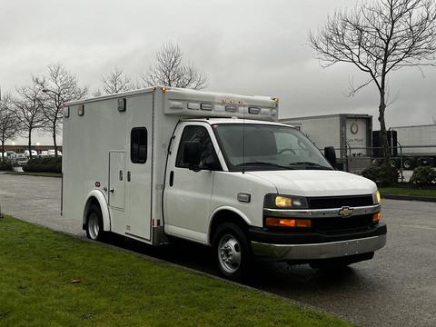 A white 2016 Chevrolet Express with a boxy cargo area and emergency lights on the roof parked on a wet surface