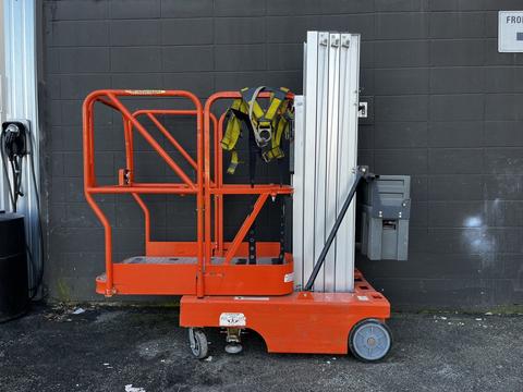 A 2000 JLG 12SP lift with a bright orange frame and aluminum platform stands on wheels with safety harness hanging on it