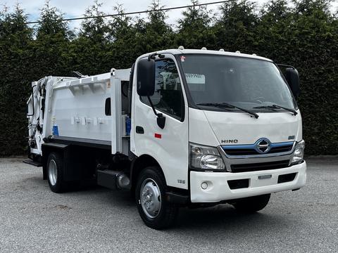 A white 2014 Hino 195 garbage truck with a closed rear compartment and side loading features parked on a gravel surface