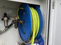A blue hose reel with yellow hose coiled inside mounted on a wall with metal fittings and an air regulator attached