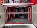 A red fire truck compartment features metal shelving with a fan and various equipment including a bell and a tool on the bottom shelf