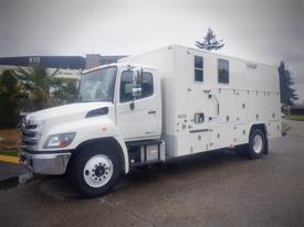 A white 2014 Hino 308 truck with a boxy cargo area and multiple windows on the side, designed for specialized transport or mobile services