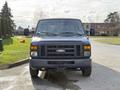 A blue 2011 Ford Econoline van is centered in the image facing forward with its headlights on and no front license plate visible