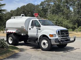 A 2015 Ford F-750 water truck with a white tank body and large wheels parked on a street
