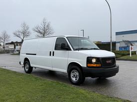 A white 2014 GMC Savana van stands on a pavement with a plain design featuring a large cargo area and front headlights illuminated