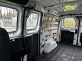 Interior of a 2023 Ford Transit showing metal framework and wooden shelving on the side walls with a grey floor and seating area visible
