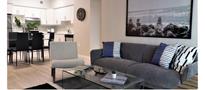 A modern living room featuring a gray sofa with decorative pillows a glass coffee table and a dining area with black chairs and a large wall art depicting a beach scene