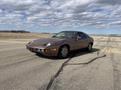 A brown 1983 Porsche 928 sports car is parked on an asphalt runway with a sleek design and distinctive headlights