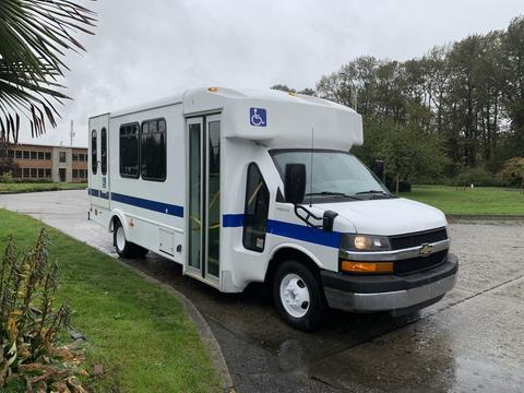 A white 2015 Chevrolet Express with a wheelchair access ramp and blue stripes on the side parked on a wet surface