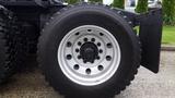 Close-up of a large truck tire mounted on a silver rim with multiple bolts and a rugged tread pattern designed for heavy-duty performance