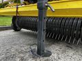 A black metal stand attached to a yellow agricultural machine with a cylindrical metal structure featuring long metallic spikes extending outward