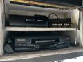 A Panasonic VHS player is displayed on a shelf with a second Panasonic device above it and various cables connected to the equipment