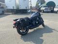 A 2020 Kawasaki Vulcan S motorcycle with a dark finish and saddle bags on the side is parked on a paved area with several trucks in the background