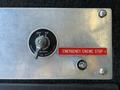 A metal control panel featuring a battery switch labeled OFF and ON along with a prominent red button reading EMERGENCY ENGINE STOP