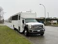 A white 2011 Ford F-550 bus with a boxy shape and large windows parked on a street along green grass