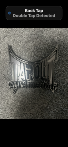 A shiny silver belt buckle with the word "TAPOUT" prominently displayed in a bold font along with the text "American" underneath