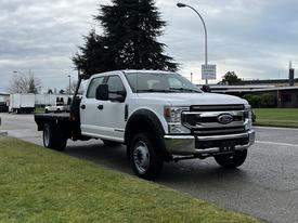 A white 2021 Ford F-550 with a flatbed design parked on the side of the road showcasing its large front grille and robust build