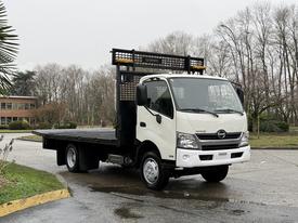 A 2015 Hino 195 flatbed truck with a white cab and black flatbed, featuring a metal mesh guard in the back and visible headlights