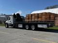 A 2012 Freightliner M2 112 flatbed truck with a hydraulic crane attachment on the rear and a large stack of wooden crates in the background