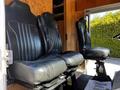 Two black leather seats designed for a vehicle, featuring vertical stitching and seatbelts, positioned side by side on a platform