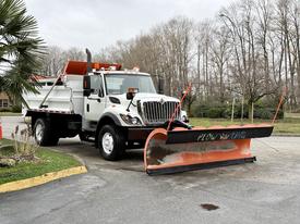 A 2012 International 7400 truck with a front snow plow attached displaying the words "PLOW" on the blade and equipped with orange warning lights