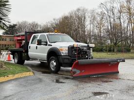 A 2013 Ford F-550 pickup truck with a snow plow attached at the front and a black hopper on the back