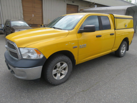 A yellow 2015 RAM 1500 pickup truck with a gray camper shell and all-terrain tires parked on a paved surface