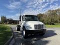 A white 2003 Freightliner M2 106 truck with a black front grill and chrome accents parked on a road with trees in the background