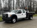 A white 2015 Ford F-550 truck with a flatbed and tool racks on the back is parked on a paved surface with trees in the background