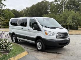 A 2017 Ford Transit T150 XLT van in white with sliding side doors and chrome accents parked at an angle