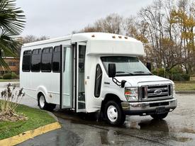 A white 2015 Ford Econoline bus with large windows and a side door, designed for passenger transport, parked at an angle with its door open