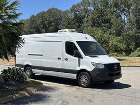 A white 2019 Mercedes-Benz Sprinter van is parked with a high roof and black wheels, showcasing a smooth design and the iconic Mercedes-Benz logo on the front grille