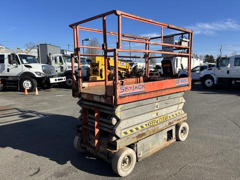 A 2010 SkyJack SJ scissor lift with an orange frame and safety railings positioned on four wheels with a warning sign that says KEEP CLEAR