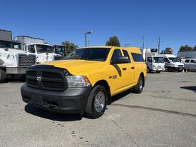 A yellow 2015 RAM 1500 pickup truck with a black grille and an enclosed bed cap parked in an industrial area