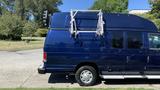 A blue 2013 Ford Econoline van with a roof rack and ladder mounted on top parked on a street